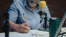 Senior man in headphones sitting at desk at studio, speaking into mic and making notes on paper when recording audio podcast