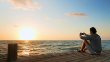 a man sitting on a dock at sunset and taking a picture with his cellphone 