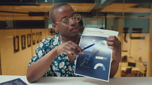 Young Black businesswoman sitting in office with glass walls and explaining financial charts on paper slide during online web conference
