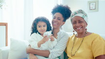 Joyous beautiful African-American woman embracing little daughter, sitting by grandmother on sofa, smiling and laughing on camera. Video portrait of happy family at home
