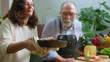 Senior couple of food bloggers taking off pan with hot stew, enjoying its smell and talking on camera, showing chefs kiss and thumbs up during online cooking class
