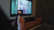 Mans hand selects internet tv channels with remote control. Close up view. Blurry tv in the background
