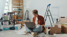 Young woman sitting cross legged on the floor among paint buckets and boxes, searching home renovation ideas on the Internet and looking around. Zoom Shot
