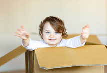 Cute smiling toddler baby girl sitting inside brown cardboard box.
