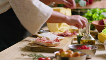 Hands of woman in apron standing by kitchen table and making gourmet sandwich with ciabatta bread, prosciutto and blue cheese. Close-up shot
