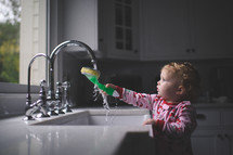 a toddler girl wetting a sponge in a kitchen sink 