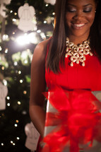 An African-American woman standing in front of a Christmas tree holding a wrapped gift 