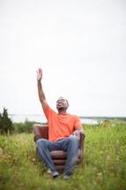 man sitting in a chair in a field with his hand raised to God 
