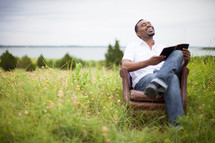 man sitting outdoors in a chair in a field reading a Bible 