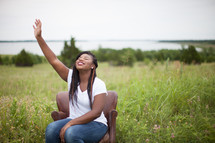woman sitting in a chair outdoors with hand raised to God 