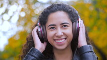 Latin curly haired girl listening music in headphones in autumn park. Beautiful young hispanic lady smiling, singing and enjoying melody outdoor.