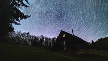 Time lapse of milky way galaxy star trails rotates over old wooden hut in forest night sky.