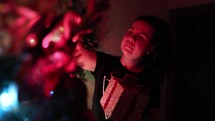 Young Woman Standing Putting Golden Gift Box On Christmas Tree. - Selective Focus