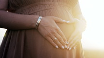 Pregnant Woman Showing Heart-Shaped Symbol Gesture On Belly, Sunlight Background. Expectant mother in linen summer dress, baby bump, happy motherhood. High quality 4k footage