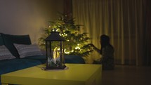 A Candle Lantern on the Table Near the Christmas Tree With a Girl Next to it - Static Shot