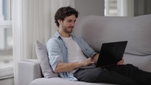 Relaxed, serious, millennial, caucasian guy using laptop device leaning on sofa at home office.
