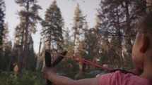 Young girl shooting a slingshot in the forest. - 2 of 2