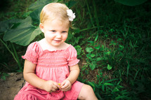Toddler girl in pink dress with flower in hair sitting in garden.