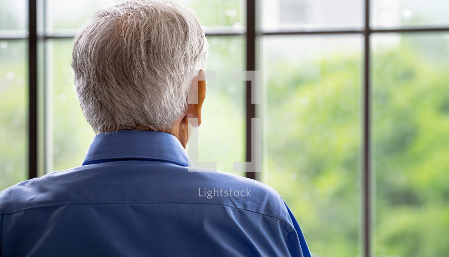 gray-haired man with back to the camera is looking out a window - suggesting waiting, loneliness, anticipation