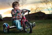 Toddler boy riding red tricycle on grass with mountains and trees in background at sunset.