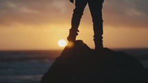 Person standing on a rock facing the sun and ocean during sunset.
