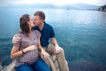 man and pregnant woman kissing sitting on a rock in front of the ocean