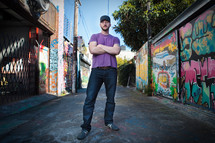 Man in jeans with purple shirt and baseball cap standing in grafitti-lined alley.