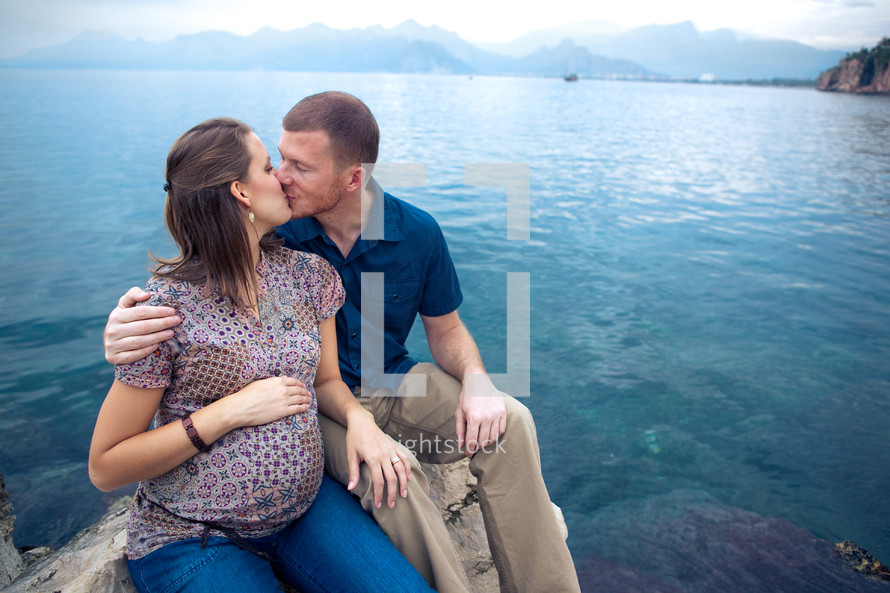 man and pregnant woman kissing sitting on a rock in front of the ocean