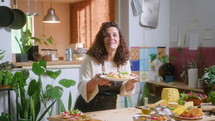 Mature female food blogger in apron putting fresh basil leaves on top of served pasta, taking plate and enjoying the smell, commenting and posing on camera with a smile in the kitchen
