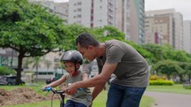 Happy latin father helping smiling boy to ride bike in city park. 
