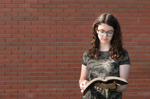 a teen girl reading a Bible in front of a brick wall 