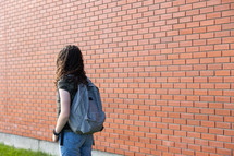 a teen girl with a bookbag standing in front of a brick wall 