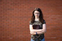 a teen girl holding a Bible standing in front of a brick wall 