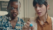 Young Latin businesswoman drawing graph on glass wall in the office and discussing financial strategy with African American female colleague
