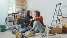 Young diverse couple sitting on the floor in the living room under renovation, looking for interior design ideas on the Internet and having discussion. Zoom Shot
