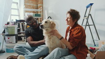 Young diverse couple petting adorable golden retriever dog and smiling when sitting on the floor among boxes, ladder and paint buckets in room under renovation
