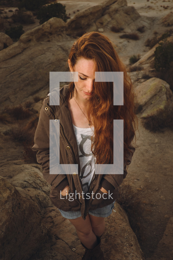 woman with long hair sitting on rocks with her hands in her pockets looking down 