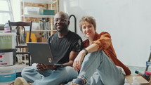 Cheerful diverse family couple sitting with laptop on the floor in the living room under renovation, posing together at camera and happily smiling. Zoom shot, video portrait
