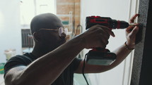 African American man in stained t-shirt and glasses drilling screw into door frame when doing home renovation or repairment
