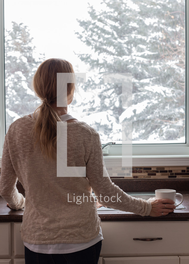 a woman looking out a window in a kitchen watching falling snow 