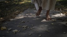 The feet of Jesus Christ or spiritual, angelic figure in white, tattered robe walking in dramatic, cinematic slow motion.