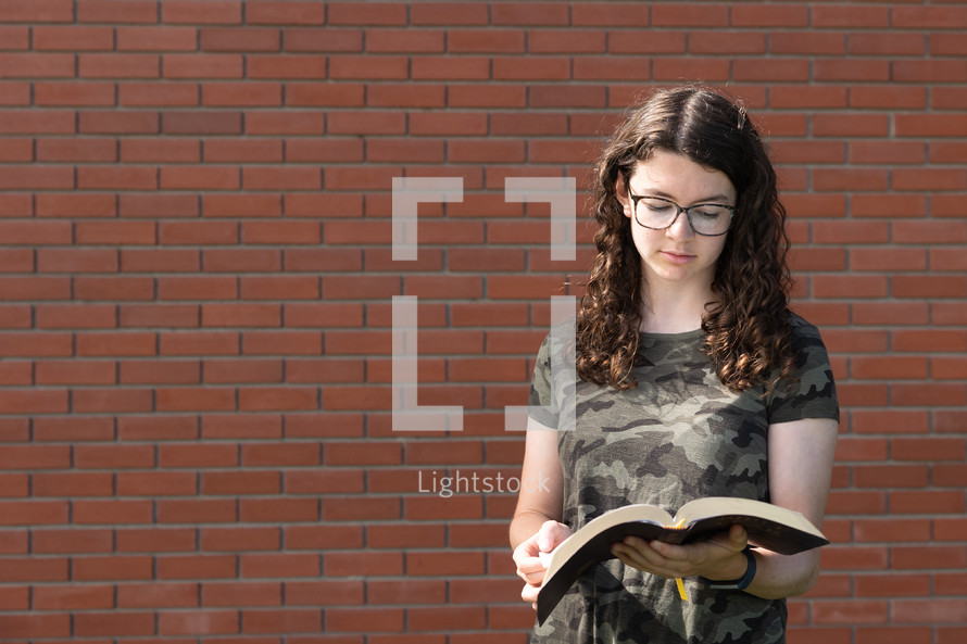 a teen girl reading a Bible in front of a brick wall 
