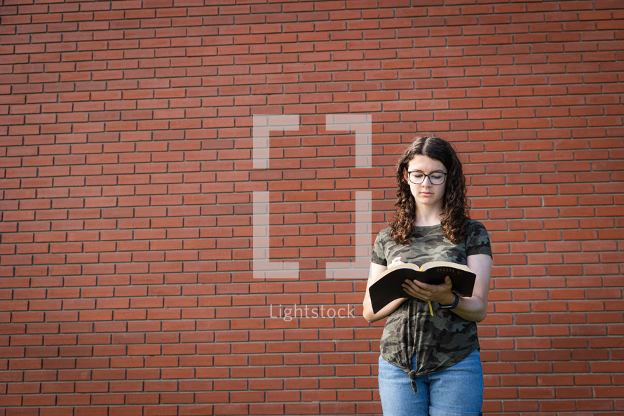 a teen girl reading a Bible standing in front of a brick wall 