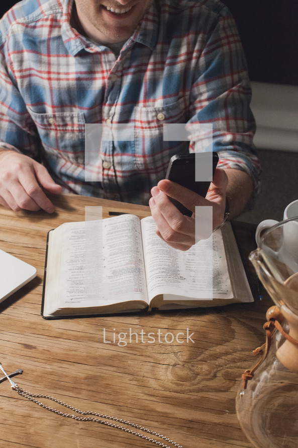 Man at a table with an open Bible, smiling and looking at his cell phone.