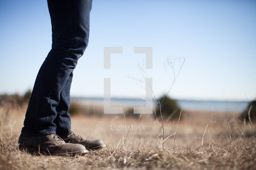 A man's legs in jeans and boots stands in a field of brown grass.