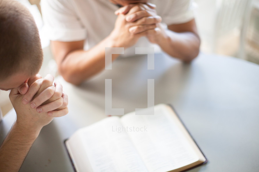 Men praying at a table over an open Bible.