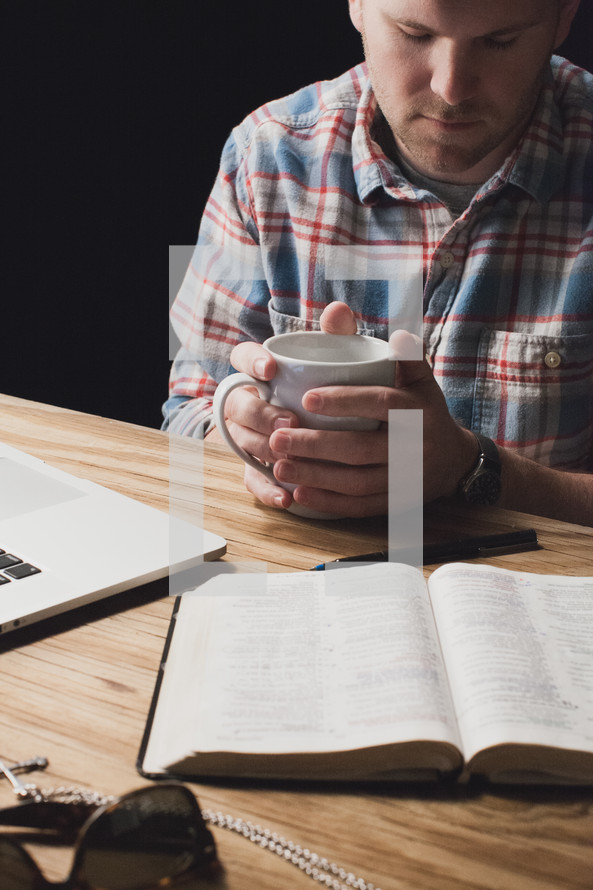 Man holding a coffee cup in prayer over an open Bible on a wooden table.