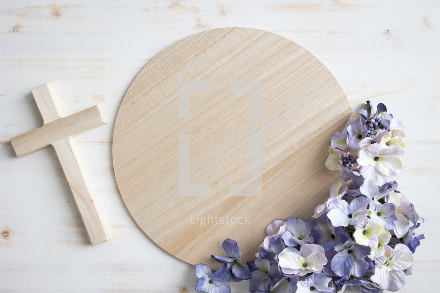 Round light wood board with frame of purple silk flowers and wood cross on a white background