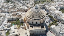 A wide view, aerial 4k drone footage, circling the Mosta Rotunda Dome, a Roman Catholic church, and the surrounding city blocks of Malta.