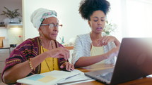 Senior black woman in headwrap and glasses sitting with papers at table, using laptop and speaking with young daughter, paying bills together online during the day at home. Medium shot
